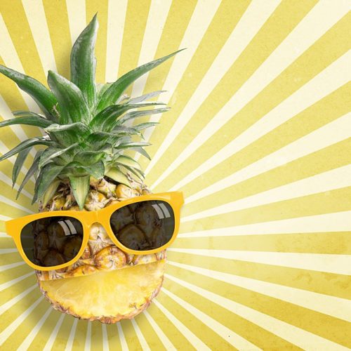 Pineapple with a cut for a smile wearing sunglasses on a background made of alternating yellow and white starburst pattern