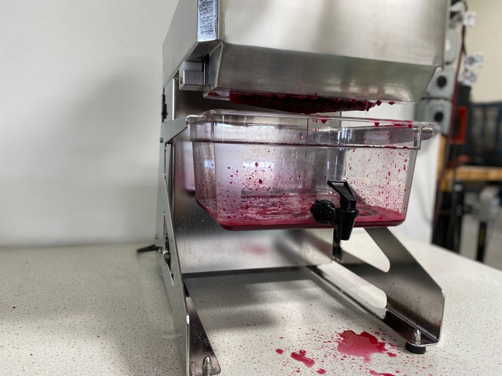 A Juiced Rite CM25 commercial cold press juicer with a juice pan with red juice in it. There's a small juice splatter by the juice pan spigot. 